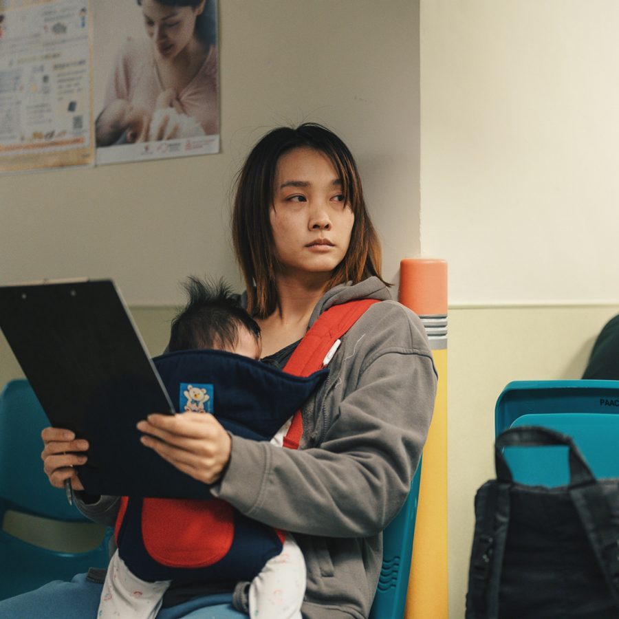 A woman sits in a medical waiting room with a newborn baby in a front carrier, in Montages of a Modern Motherhood