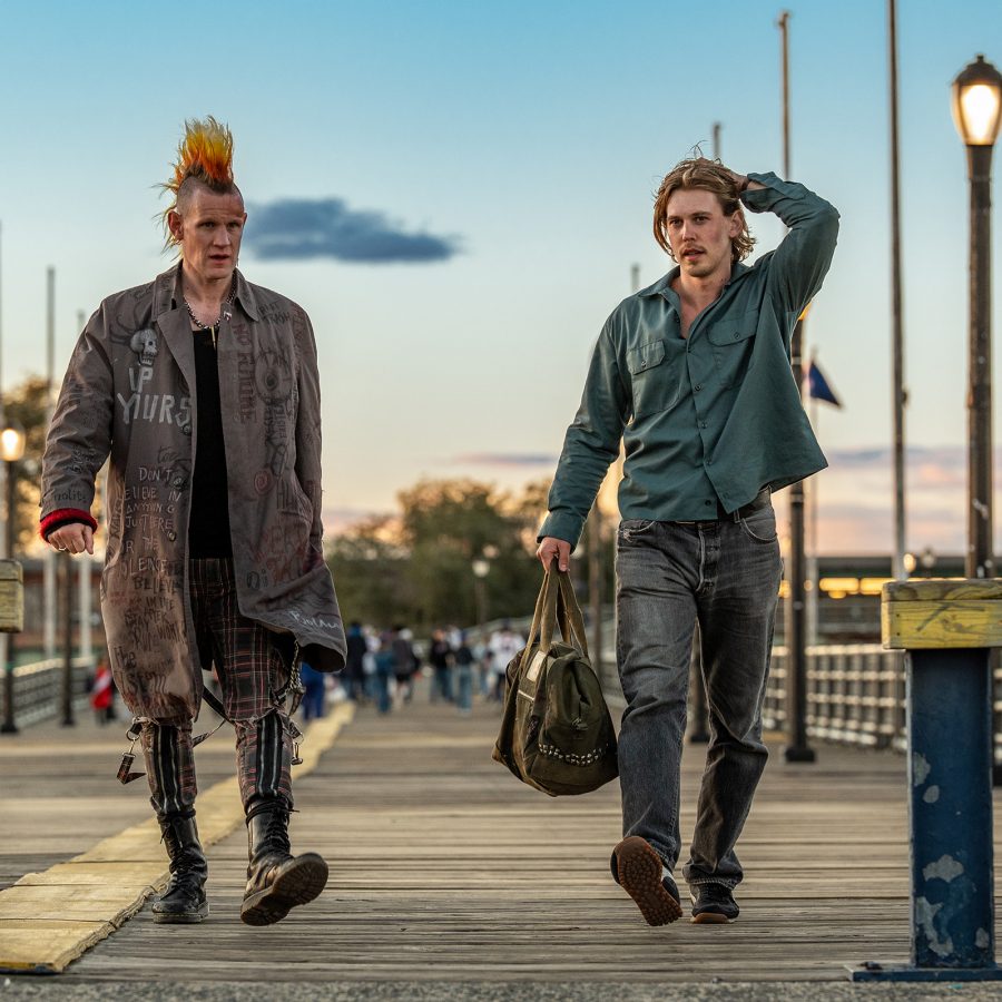 Two men – one dressed as a punk with a mohawk haircut, the other in casual clothing – walk along a pier at sunset in a scene from Caught Stealing