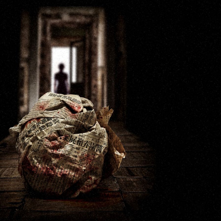 A scene from She’s Got No Name – in a dark room, light shines on a ball of newspaper that is stained with blood; behind it, a woman’s silhouette can be seen in a doorway.