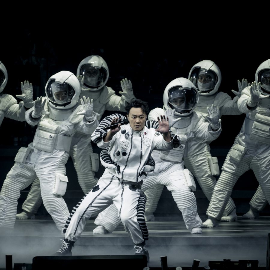 Dressed in white and black, singer Eason Chan performs on a stage with a group of dancers who are dressed as astronauts, in a scene from FEAR and DREAMS: NOW is the only reality.