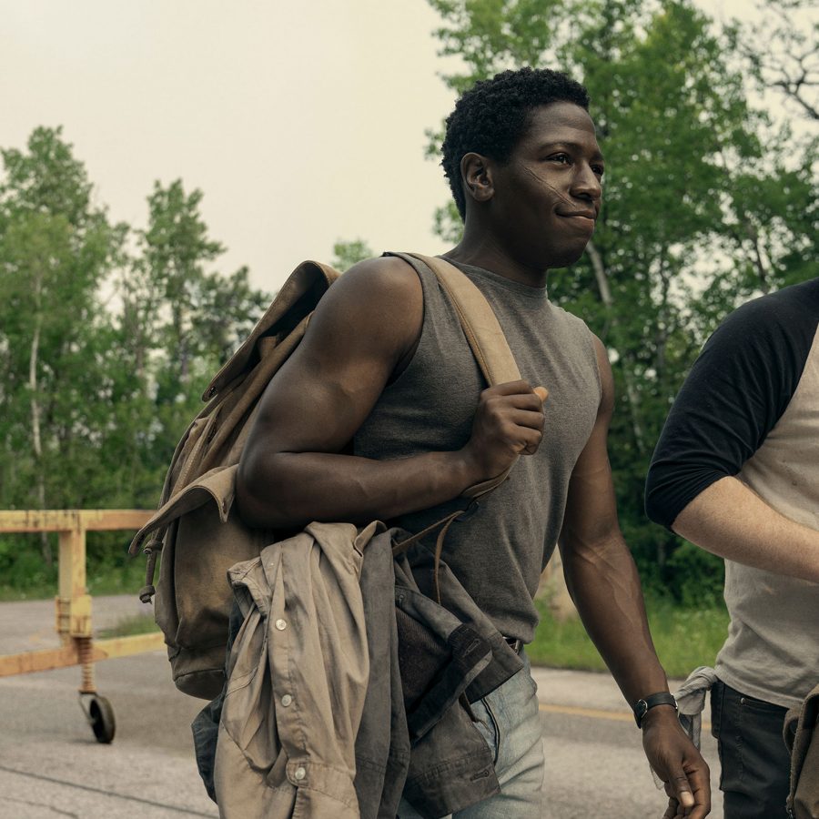 Two teenage boys carrying rucksacks walk along a remote road, while an armed military officer stands behind them, in a scene from The Long Walk