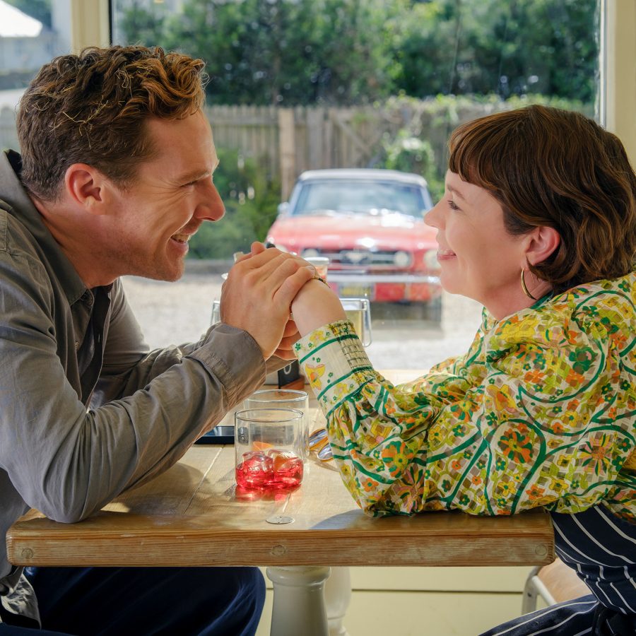 A man and woman sit a restaurant table, holding hands and looking lovingly at one another, in a scene from The Roses.