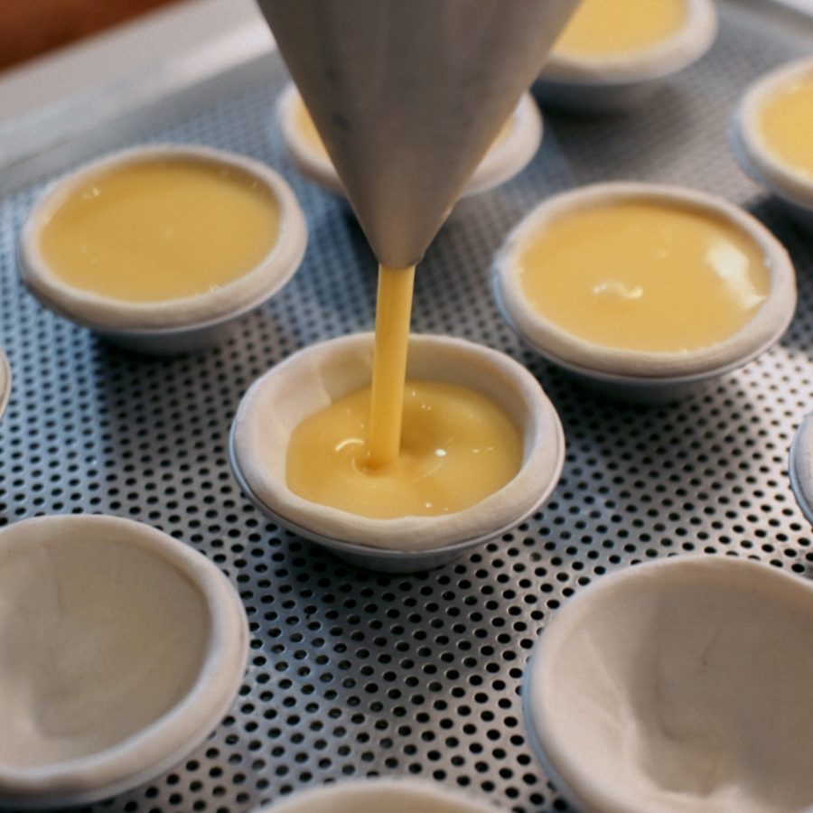 Custard is piped into egg tart shells lined up on a metal baking tray. 