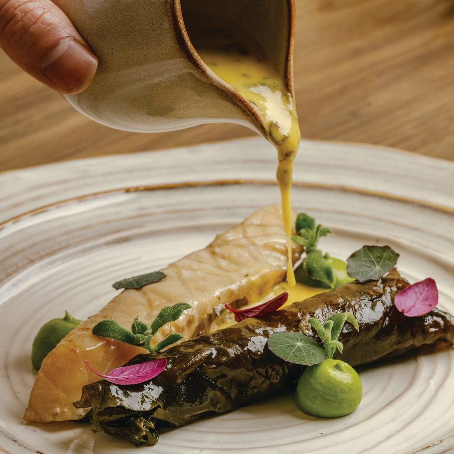 Plated dish at Racines in Hong Kong, featuring a fillet of fish served alongside leaf-wrapped vegetables, green purée and edible flower petals. A sauce is being poured from a pitcher onto the dish on a cream-coloured plate.