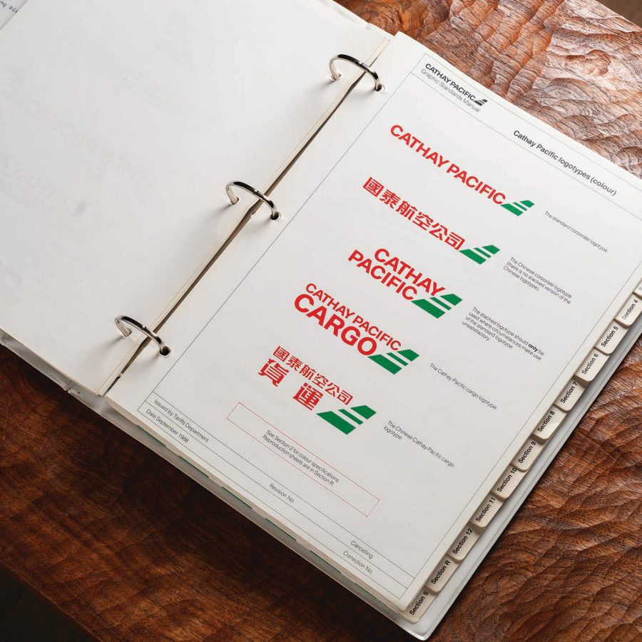 Open ring binder with tabbed sections, open to a page with historical Cathay Pacific logos in red and green, displayed on a wooden tabletop.