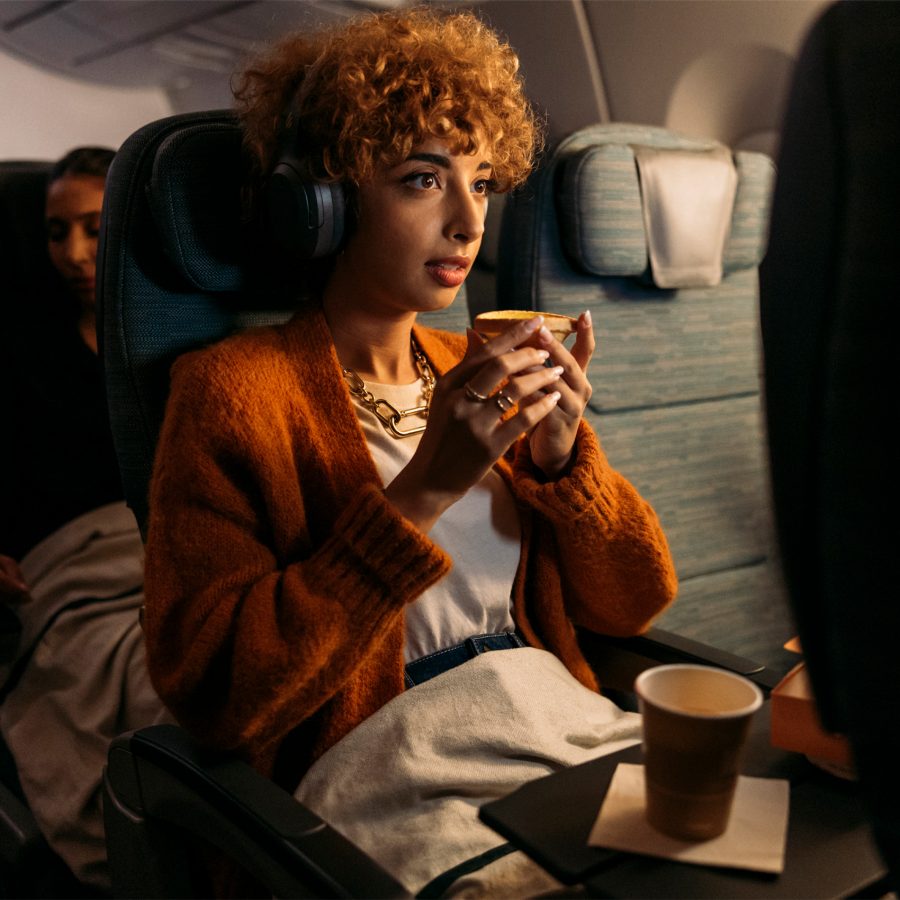 A lady with curly ginger hair and wearing an orange cardigan is watching the Inflight Entertainment system on a Cathay Pacific flight and holding an egg tart.