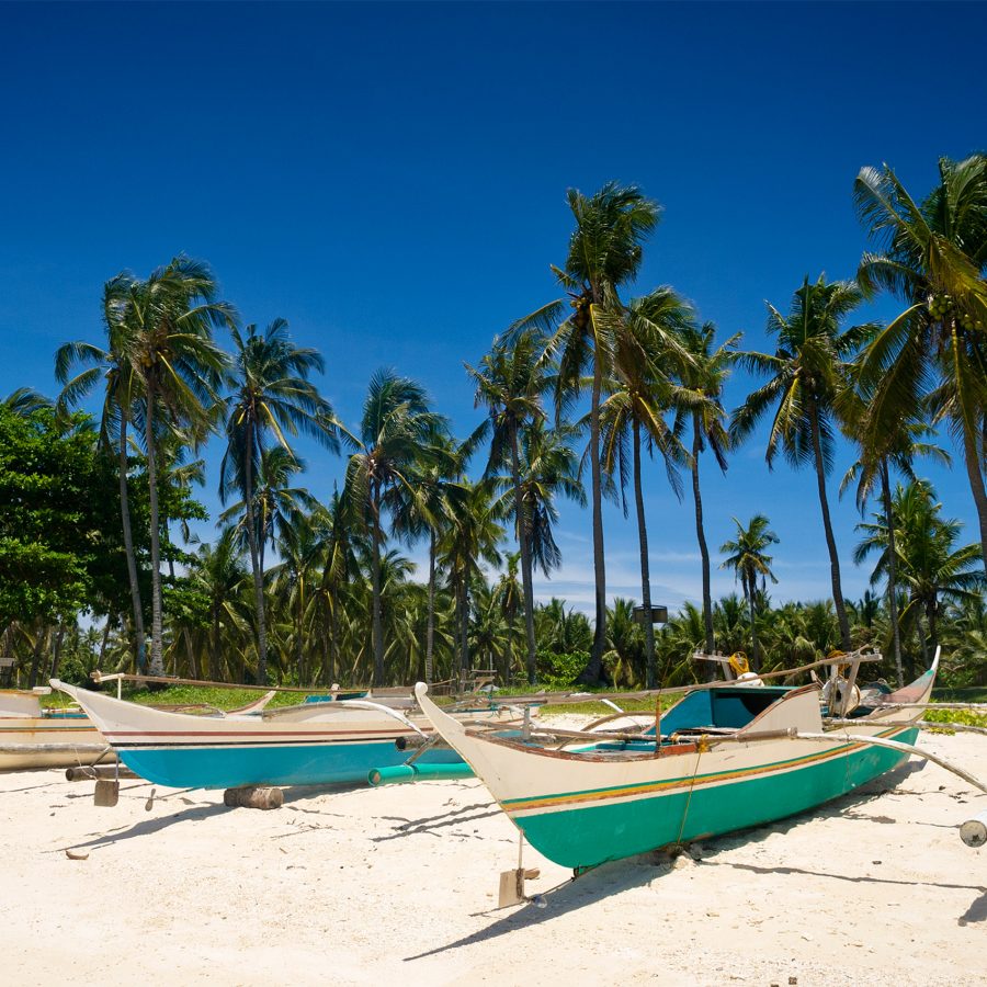 Two wooden Bangka boats parked on a white sandy beach with palm trees and clear blue sky in the background