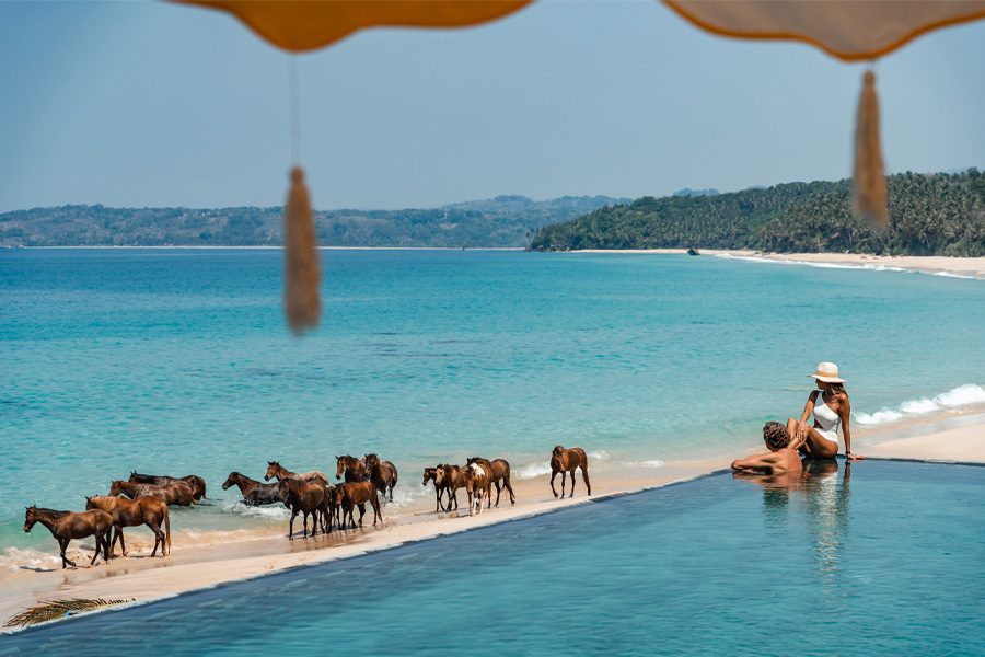 A couple enjoying a day at the pool with the view of the sea and horses on the beach at Nihi Sumba, in Sumba, Indonesia
