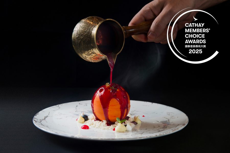 A hand pours red sauce over an edible orange ball on a plate. 