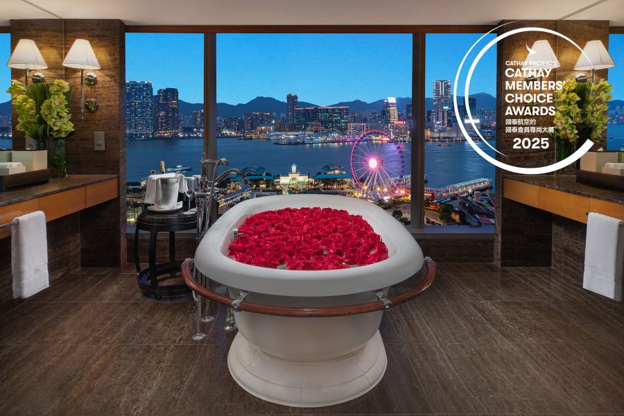In a guest room at Mandarin Oriental Hong Kong, a white freestanding bathtub is covered in rose petals and overlooks the Central Pier and the Hong Kong Observation Wheel during the evening.