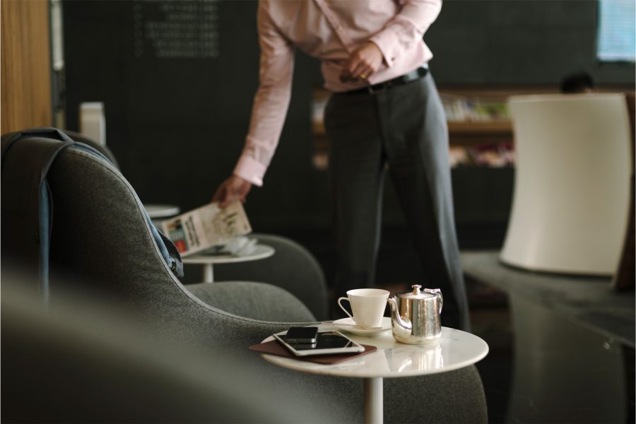 A cup and teapot sit on a marble side table alongside an iPad in a lounge, while a man puts a newspaper down at a nearby table.