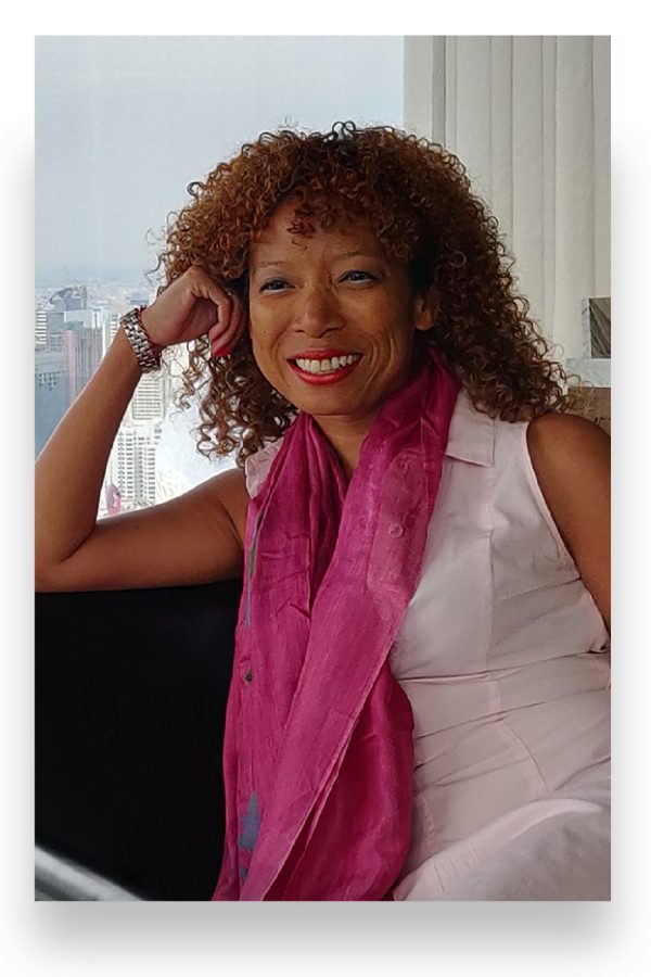 Travel+Leisure Southeast Asia Editor-in-Chief Jennine smiles as she sits in a chair with a skyline behind her through floor-to-ceiling windows.