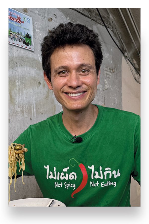 Food influencer Mark Weins smiles as he eats a bowl of noodles in a restaurant.