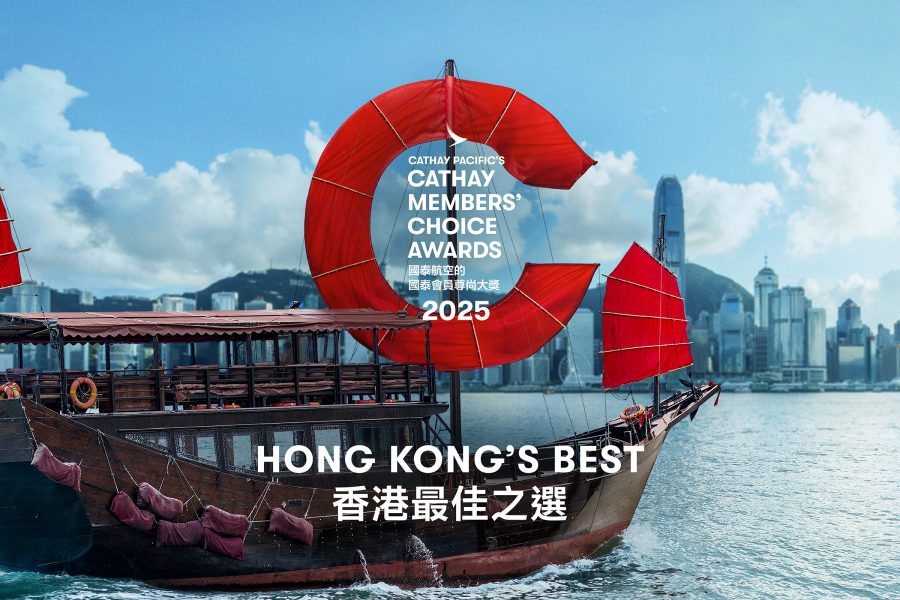A red-sail Chinese junk boat floats in Victoria Harbour in front of Hong Kong's skyline. Its sail is shaped like a C.