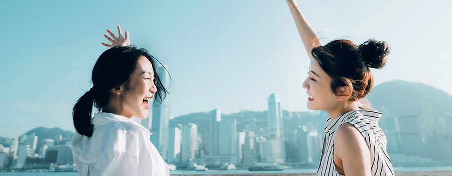 Two young women happily facing each other and waving on a waterfront promenade, with a harbor and tall buildings in the background.