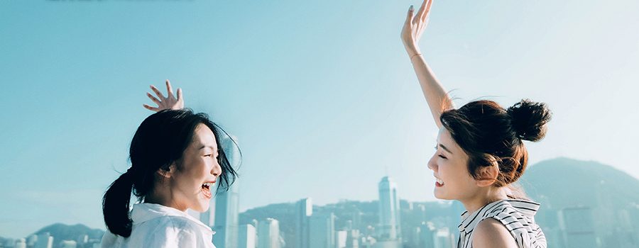 兩位年輕女子在海濱長廊上高興地對看和揮手，背景是海港和高樓大廈。