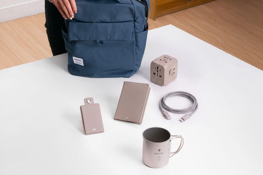 Cathay Shop travel essentials neatly arranged on a table, featuring a compact backpack, travel adapter, charging cable, luggage tag, passport holder, and mug.