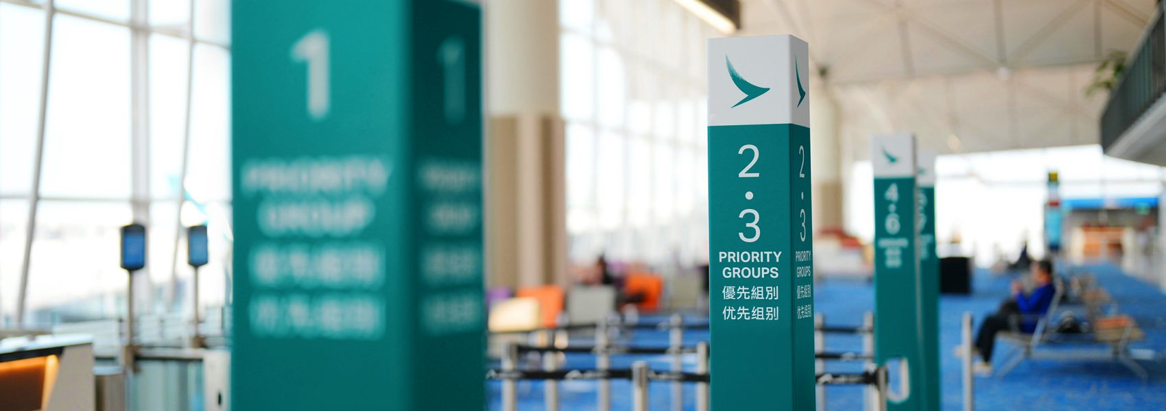 Green and white boarding group signage numbered 1, 2 and 3 stand in a row inside Hong Kong International Airport. The background shows blue carpeted seating areas and large windows letting in natural light.