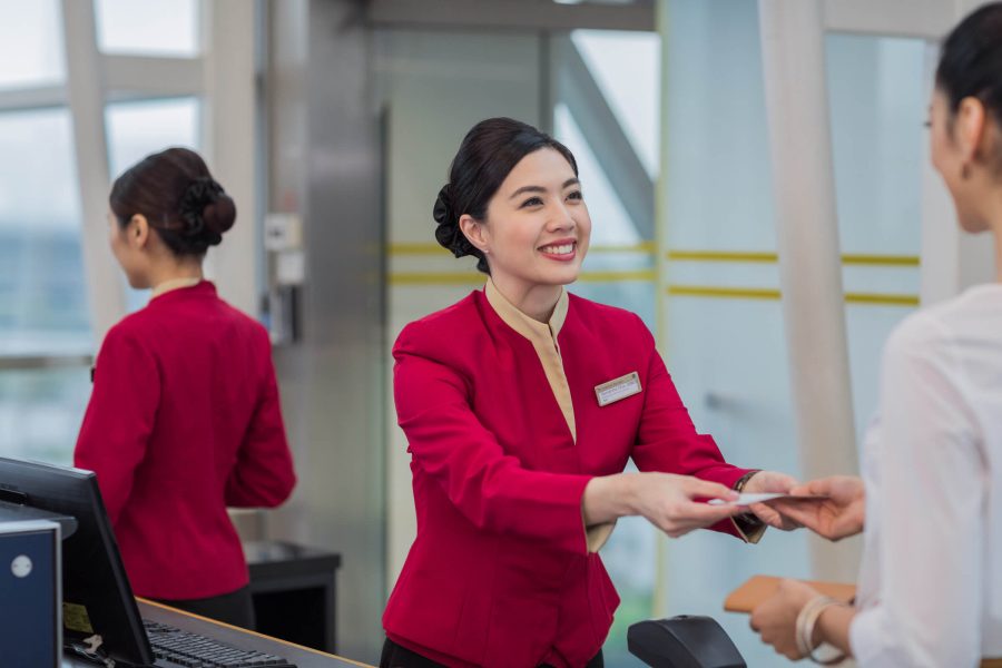A smiling staff member in a red uniform hands a document to a customer across a service counter, with another staff member in the background reflected in glass. The setting resembles an airport or service desk.