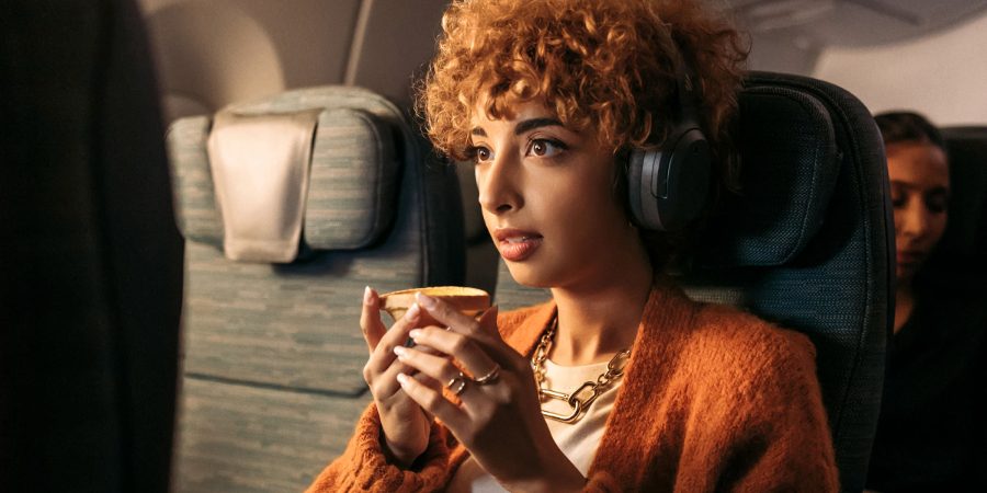 A female passenger wearing headphones is sitting in a Cathay Pacific Economy cabin and looking at the seat-back screen in front while holding a cup. 