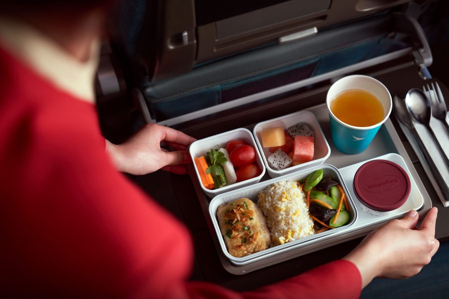 A cabin crew places an ensemble of food, including fruits, an appetiser, a main course, a cup of tea and ice cream, onto an Economy tray table.