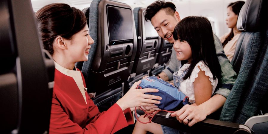 Un miembro de la tripulación de cabina de Cathay Pacific muestra a una familia tres dibujos y textos en un trozo de papel dentro de una cabina de clase Economy. El niño, sonriente y vestido de rosa, está sentado en el regazo de su madre.