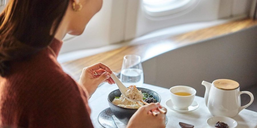 A woman eating a bowl of wonton noodles with chopsticks and spoon, with Chinese condiments, a cup of tea and a glass of water aside, in Cathay Pacific’s First class cabin.     