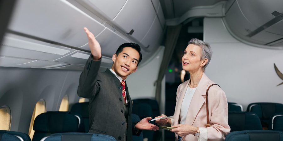 A flight attendant in a suit assists a passenger inside an airplane cabin, pointing toward the overhead compartments. The passenger, holding a boarding pass and documents, looks attentively in the indicated direction. The cabin features blue upholstered seats and overhead storage bins, suggesting a boarding or pre-takeoff moment.