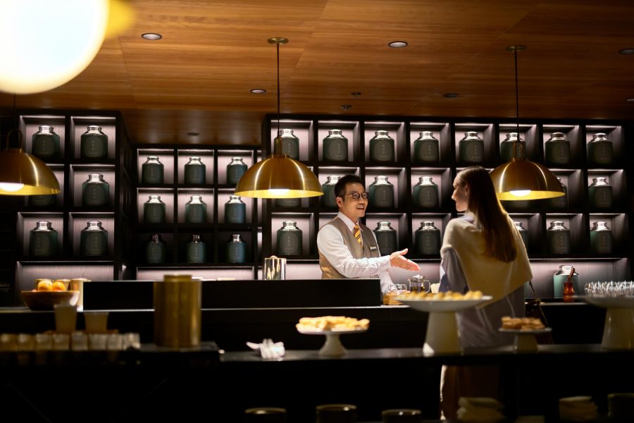 A warmly lit, upscale café or bar with a wooden ceiling and pendant lights. A male server chats with a female customer across a counter displaying pastries and glassware. Behind them, shelves lined with black-labeled jars create a stylish, organized backdrop.