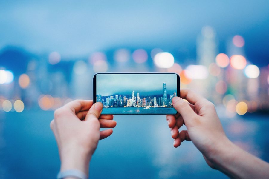 A pair of hands holds a smartphone, capturing a photo of a city skyline with tall, illuminated buildings. 