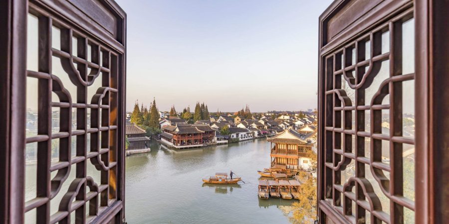 A picturesque view of a Chinese ancient town framed by ornate wooden window panels, showing wooden buildings with traditional Chinese architecture on the riverside and sampans on the calm water. 