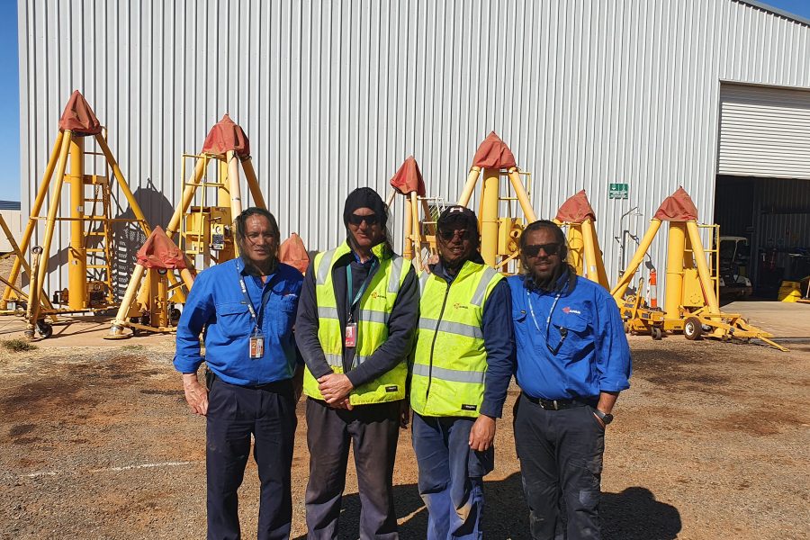 Four workers in blue uniforms and safety vests standing near yellow industrial equipment outside a warehouse.