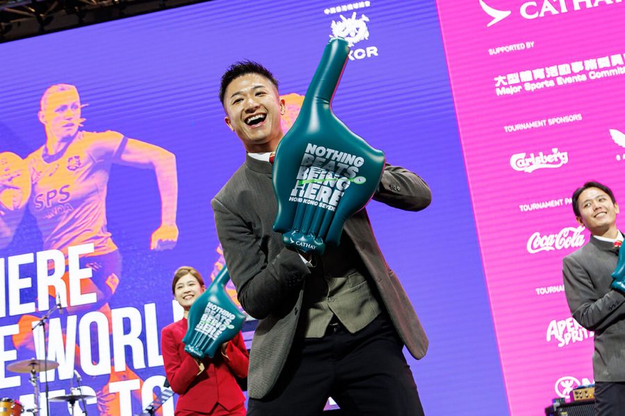People on stage holding large foam hands with the text ‘NOTHING BEATS BEING HERE,’ in front of a vibrant event backdrop featuring a rugby player graphic and the slogan ‘WHERE THE WORLD COMES TO PLAY,’ along with sponsor logos including Cathay Pacific and Coca-Cola.