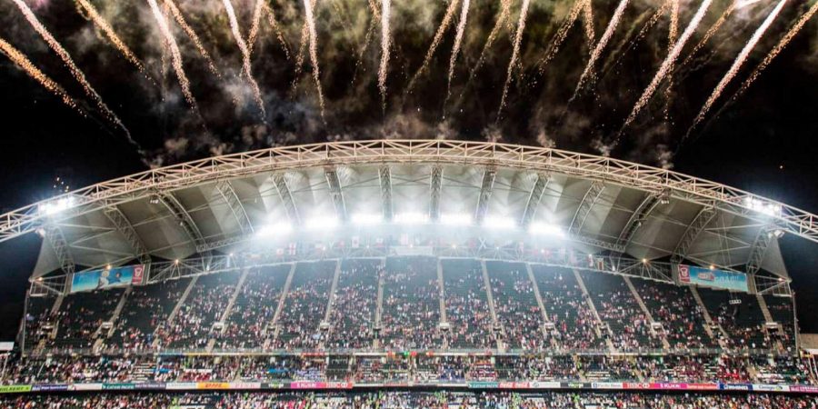Large stadium filled with spectators during a nighttime event, with bright fireworks shooting across the sky above the curved roof structure. The field is partially visible at the bottom, and colorful advertising banners line the stands