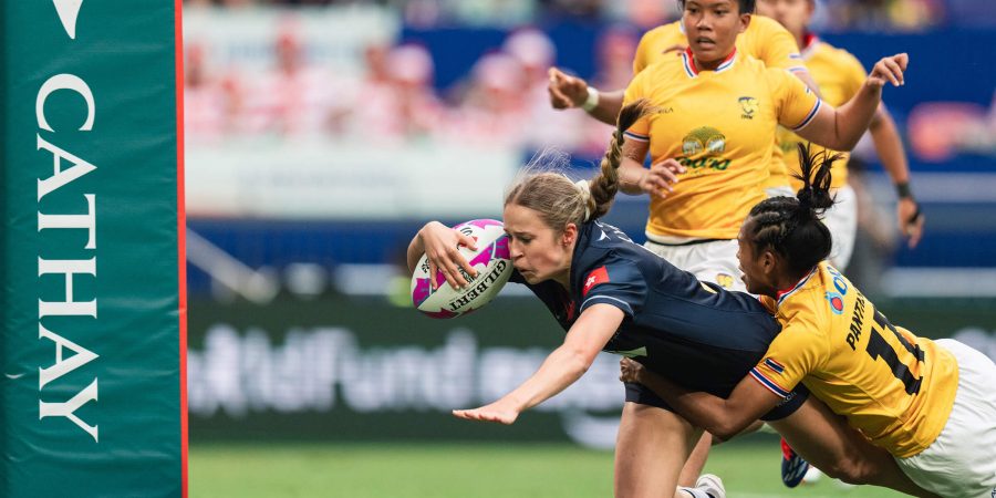 Rugby player in a navy blue jersey diving forward to score a try while holding a pink-and-white ball, tackled by an opponent in a yellow jersey near the goalpost marked with the Cathay logo. Other players in yellow jerseys are visible in the background on a green grass field.