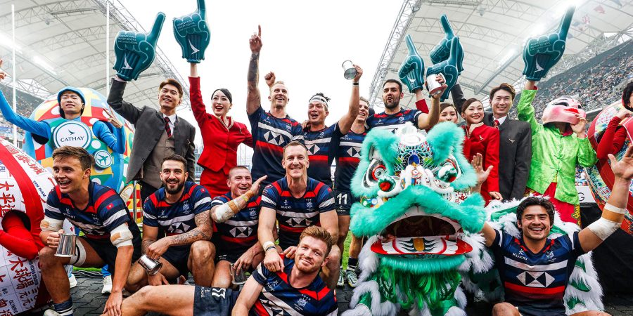 Group of rugby players in navy blue and red striped jerseys posing with trophies and foam hands, joined by people in suits and a performer in a bright green costume next to a colorful traditional lion dance figure. The scene is set in a large stadium with a covered roof and a cheering crowd in the background.