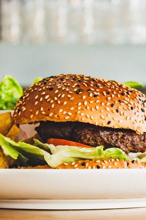 Close-up of a burger served on a white plate. The bun is speckled with black and white sesame seeds, and sandwiched inside are lettuce, tomato slices and a patty.