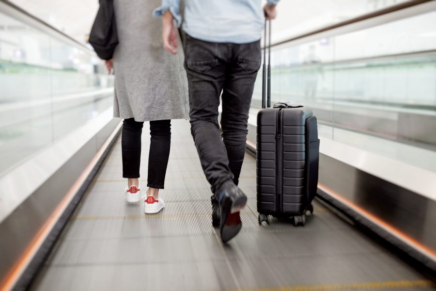 A lower-half body shot of two people on a moving walkway shot from behind. One of them is with a black carry-on baggage.