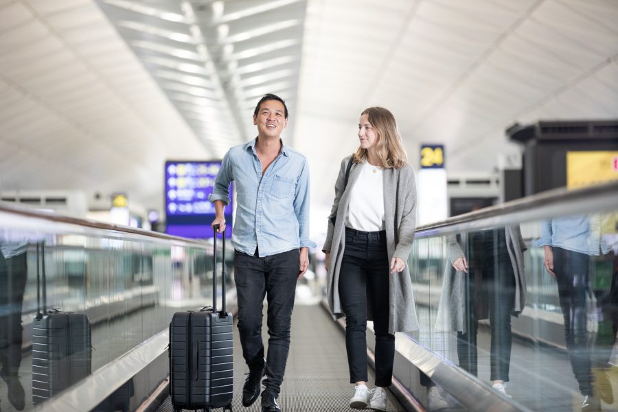 A man and a woman walking on a moving walkway in the restricted area of Hong Kong International Airport. The man has a black carry-on baggage with him.